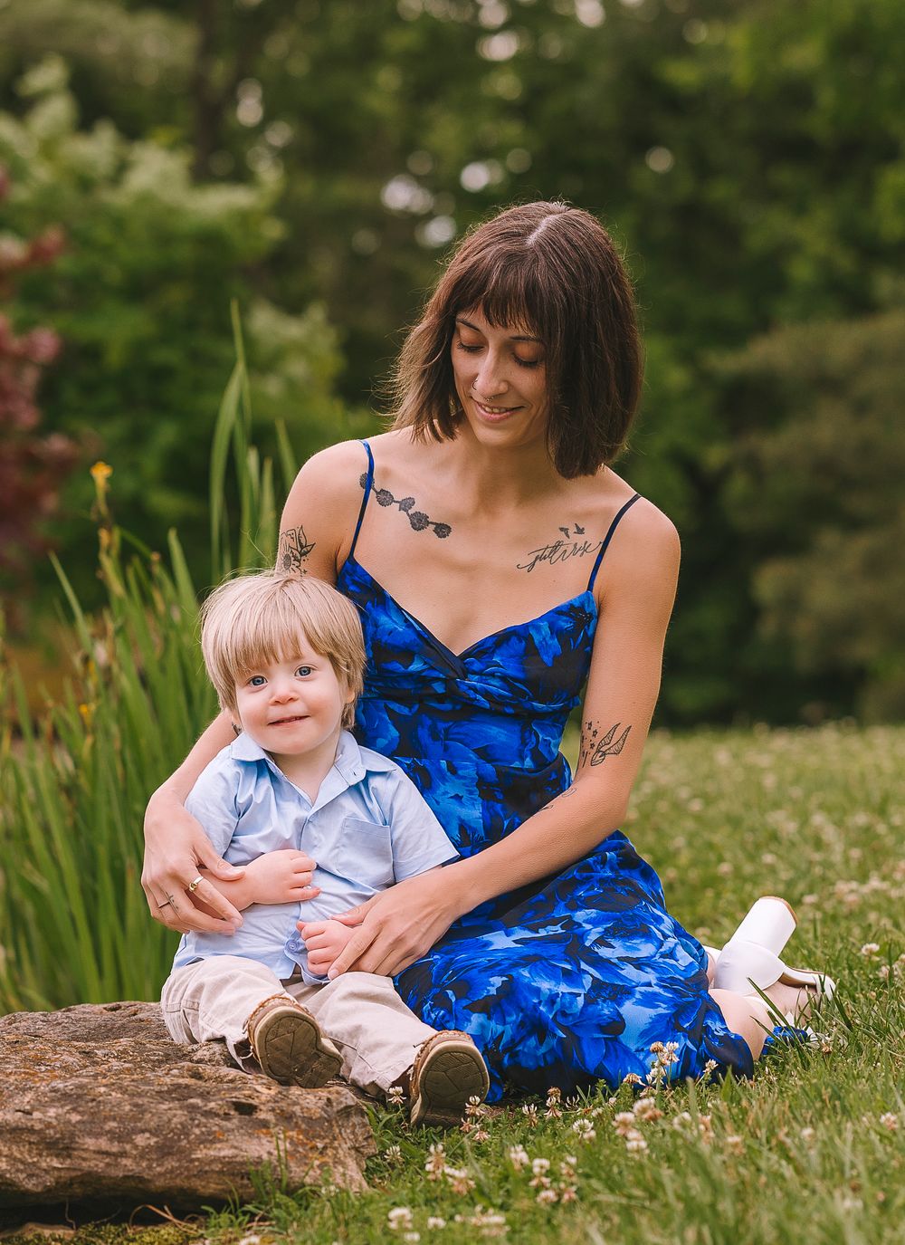 A person in a blue floral dress sits with a young child on a log in a grassy outdoor setting on a summer day.