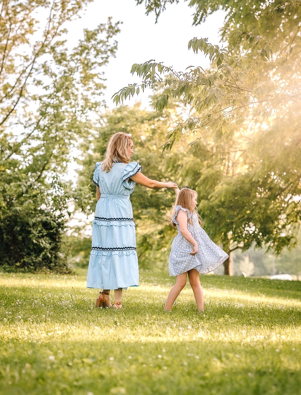 A young girl in a blue dress twirls and dances on a sunlit grassy hill surrounded by trees.