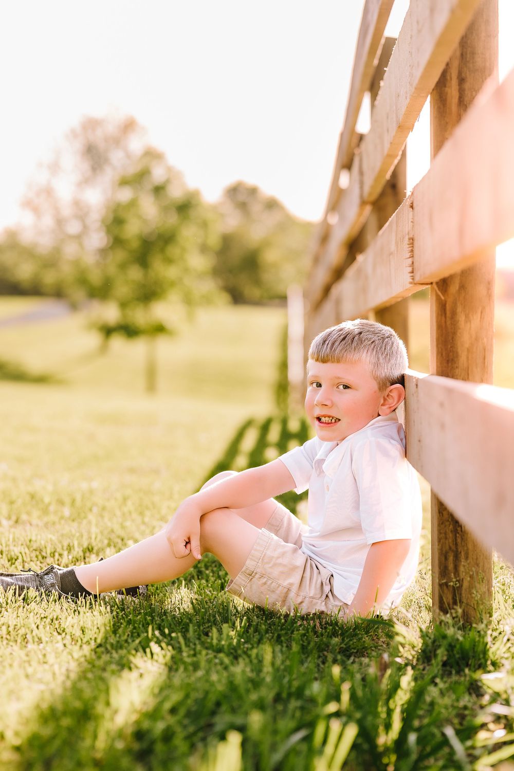 Young child sits barefoot in green grass leaning against a wooden fence rail in warm summer sunlight.