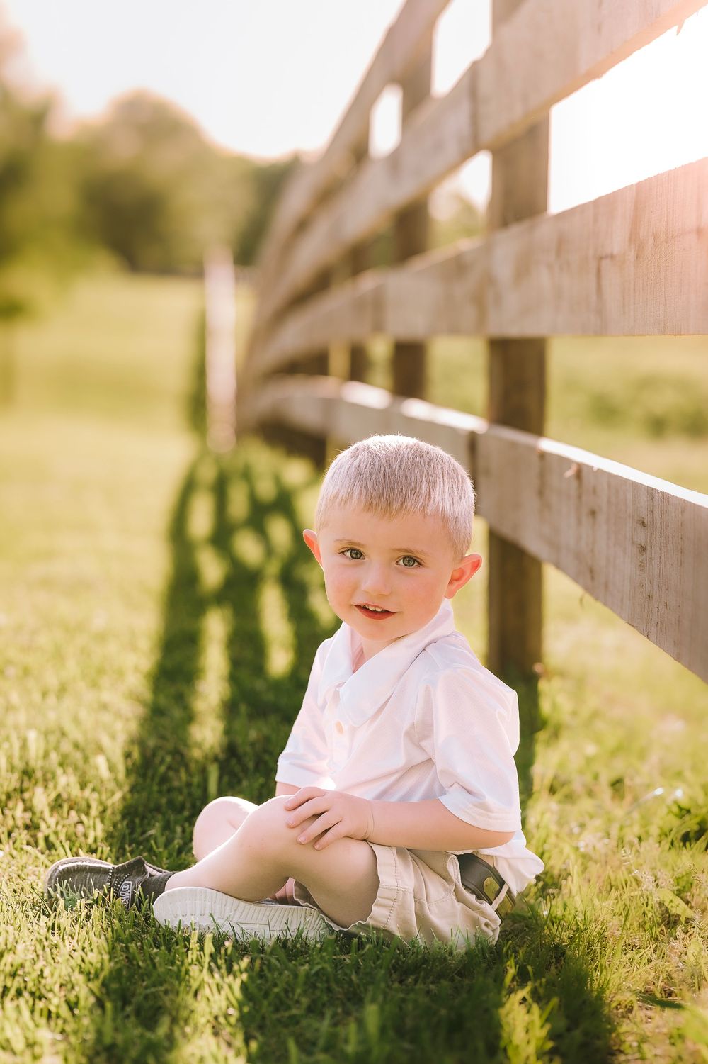 Young child sitting in soft sunlit grass beside wooden fence panels on a warm summer evening.