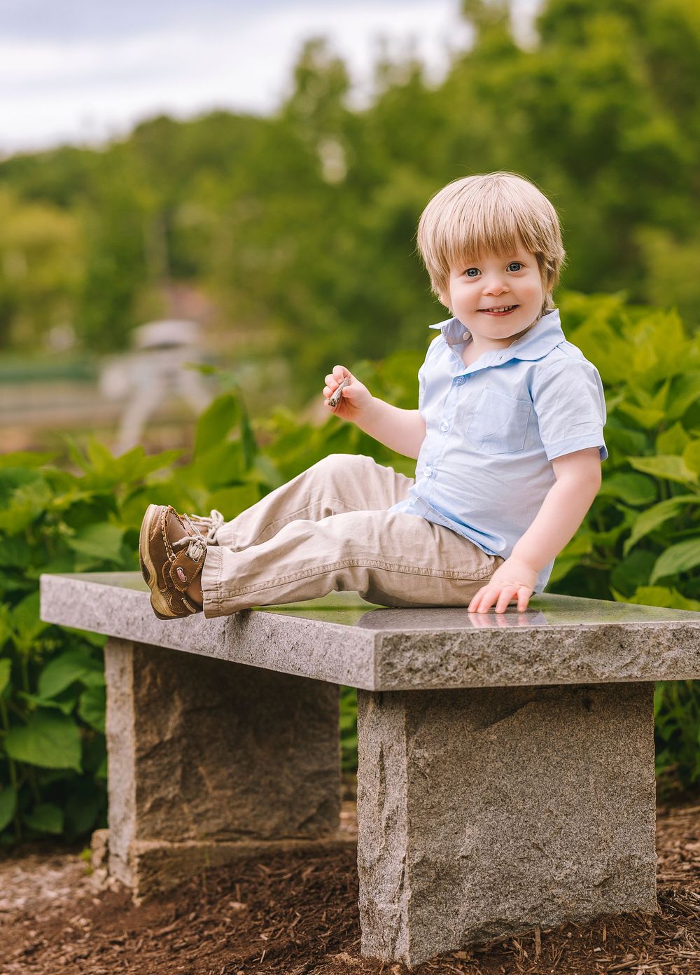 A young child poses playfully on a rustic stone bench surrounded by lush green foliage in a garden setting.