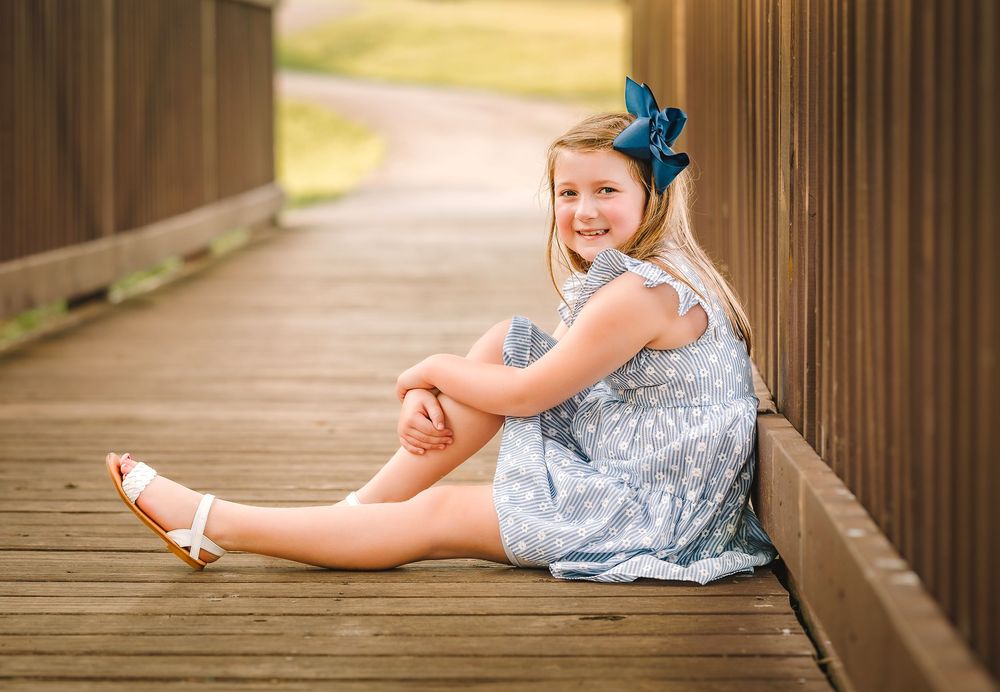 A child in a blue dress and matching hair bow sits on a wooden bridge walkway during a sunny day.