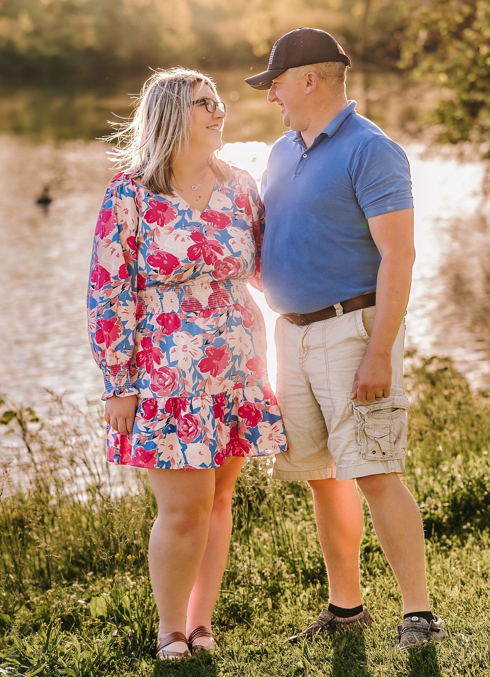 A couple stands together by a lake, with her wearing a floral dress and him in a blue polo and khaki shorts on a sunny day.