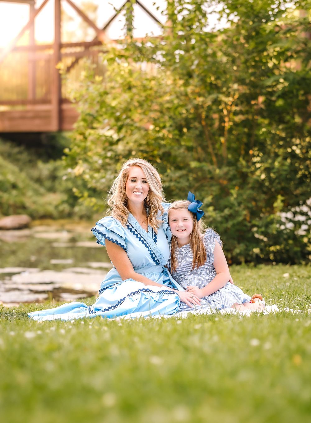 A mother and daughter in matching blue dresses sit together on a blanket in a lush garden setting during sunset.