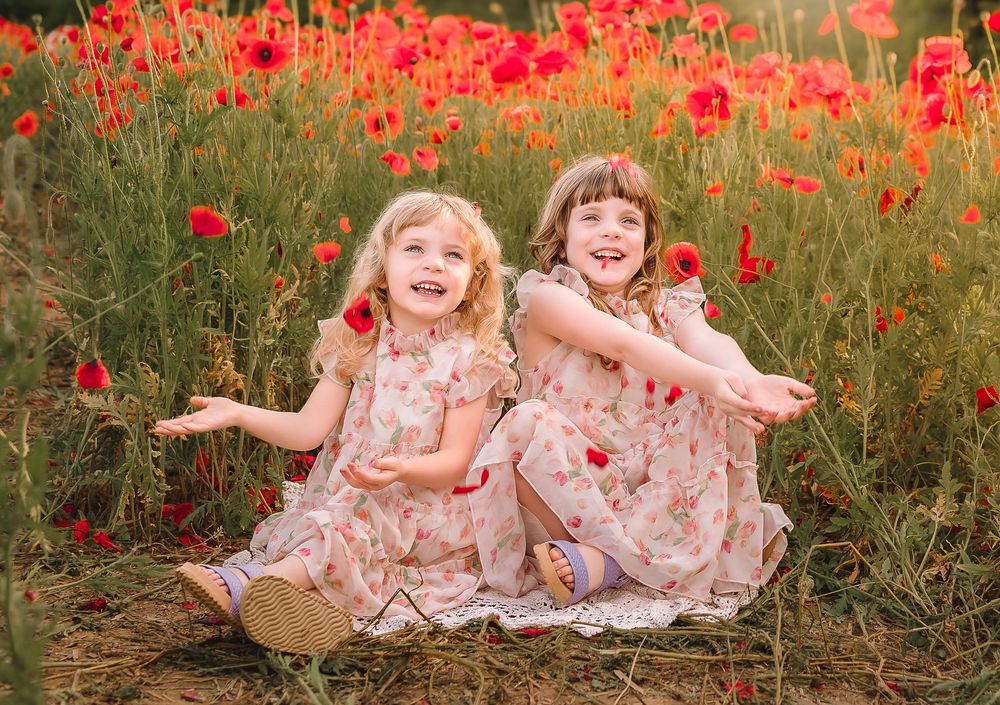 Two young children in floral dresses laughing and playing together in a vibrant red poppy field during sunset.
