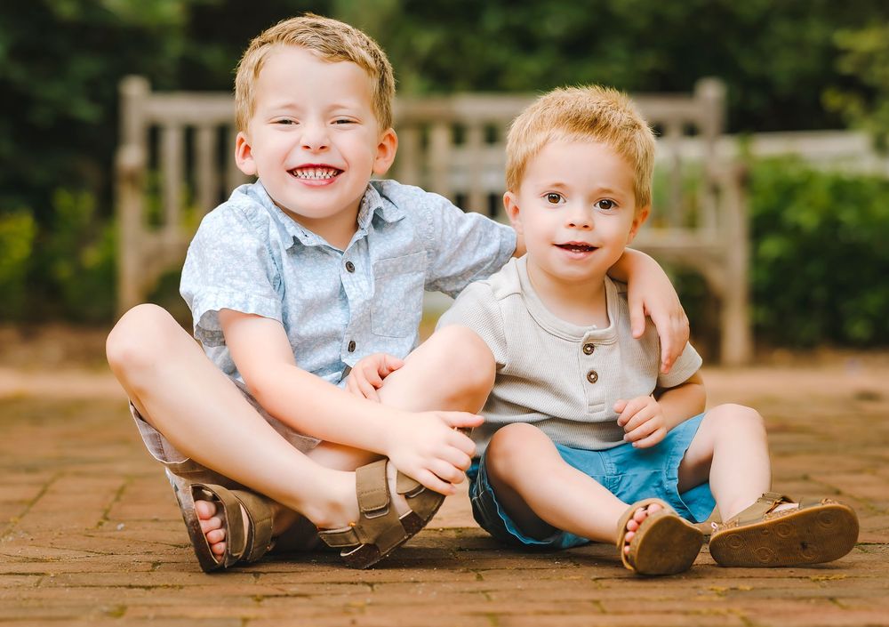 Two children sit together on a wooden deck while smiling and wearing casual summer clothing.