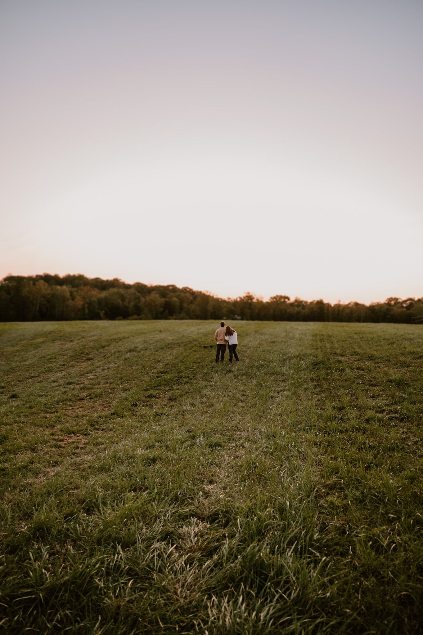 Couple stands together in vast open green field at golden hour with tree line backdrop Lexington NC