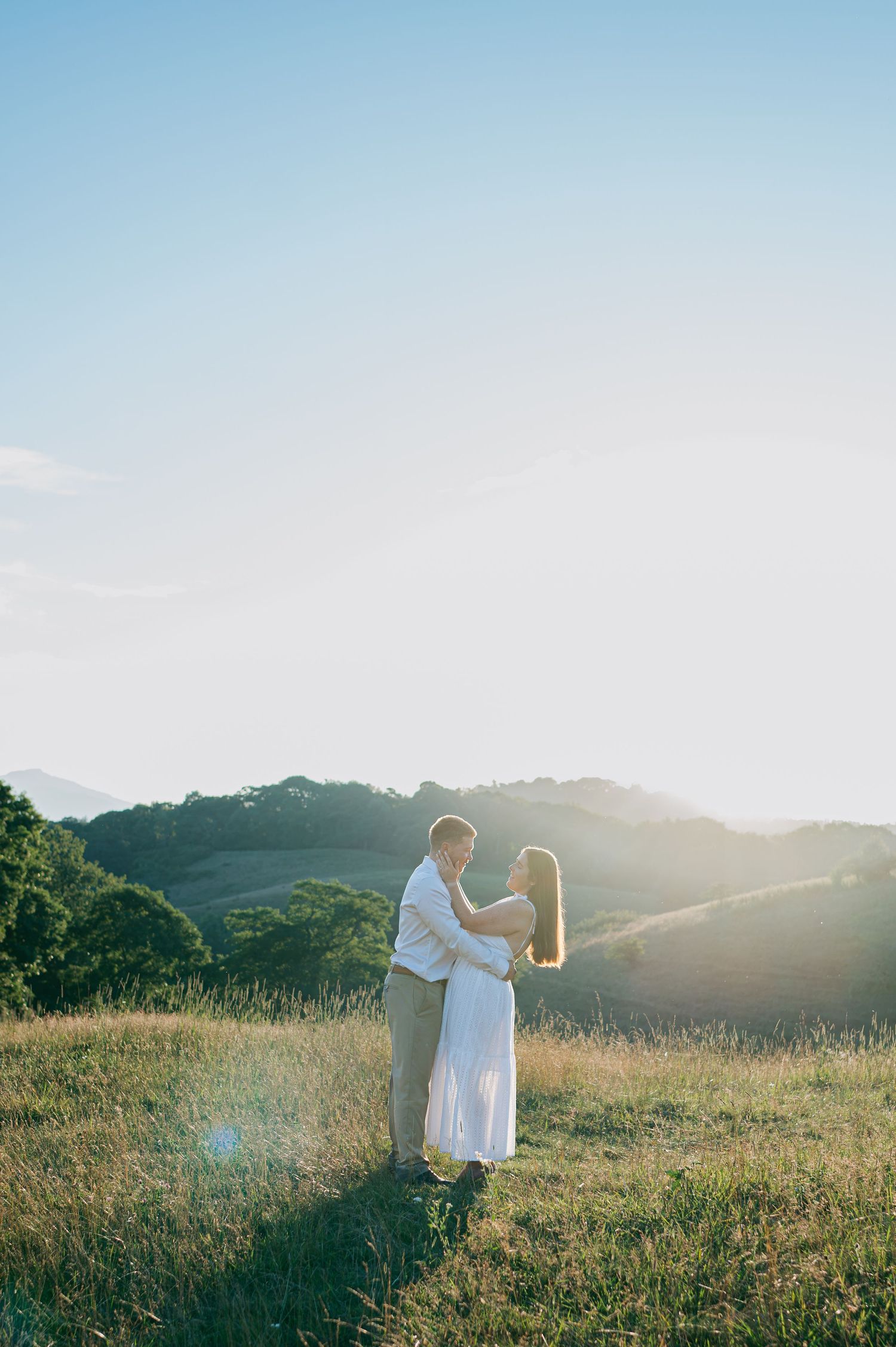 Couple embraces in sunlit golden hillside meadow with rolling green hills in background Lexington NC
