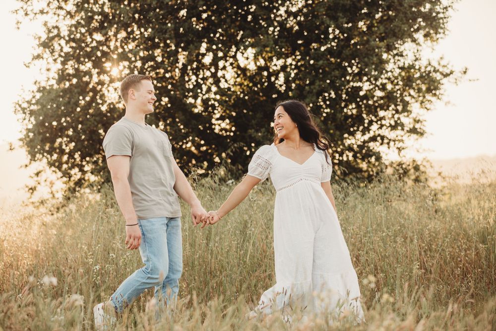 A couple holding hands in a sun-drenched field with tall grass and trees silhouetted in the golden hour light.