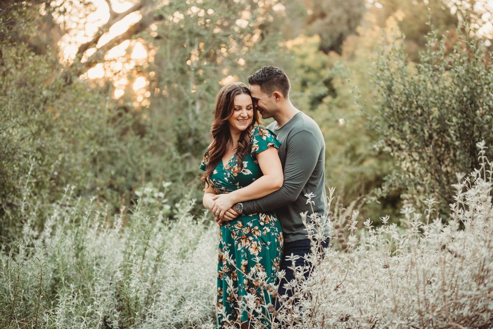 A romantic couple embraces in a sun-dappled garden setting among green foliage and delicate white wildflowers.