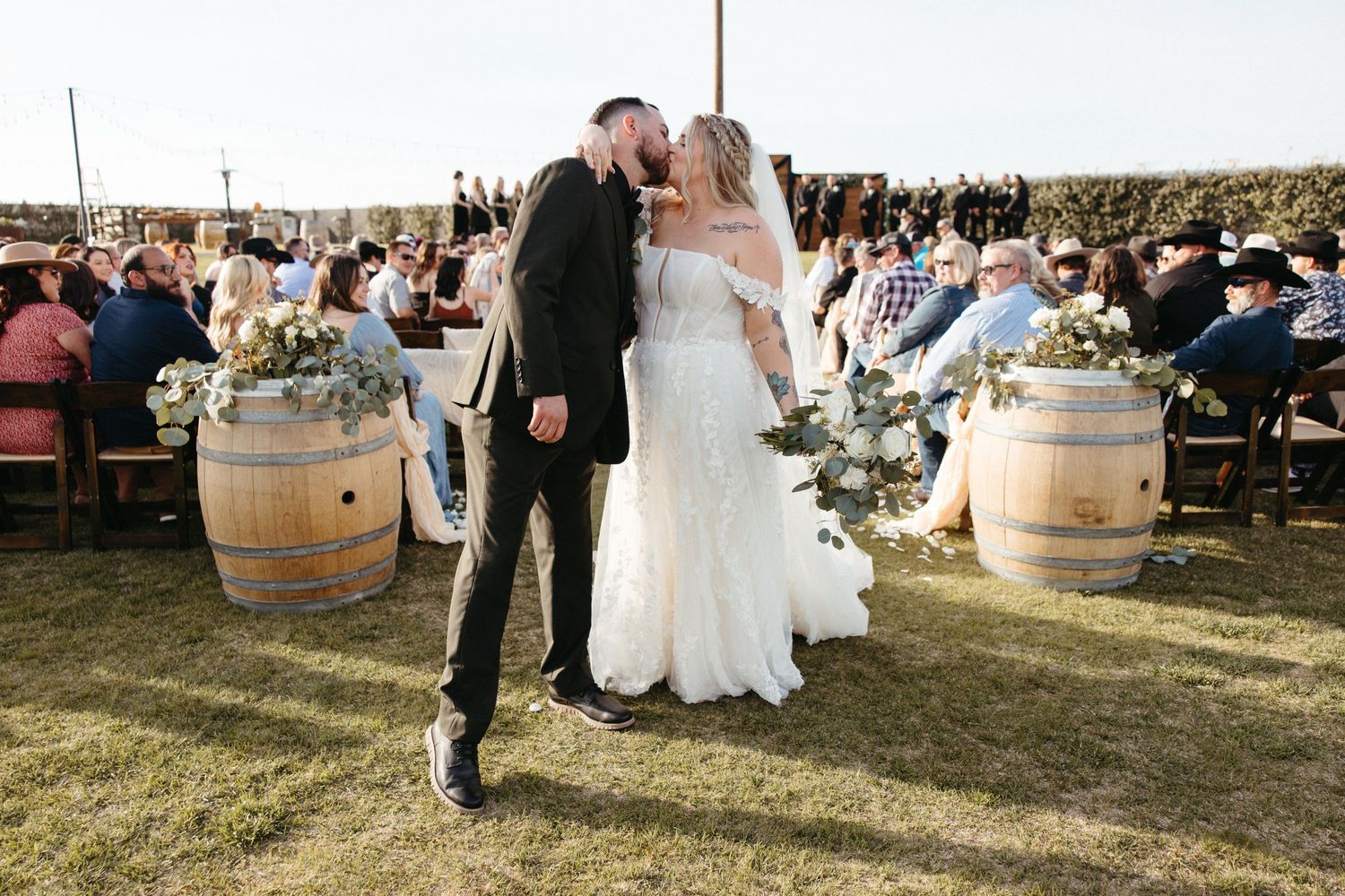 A romantic outdoor wedding ceremony with wine barrels and guests seated on a grassy field.