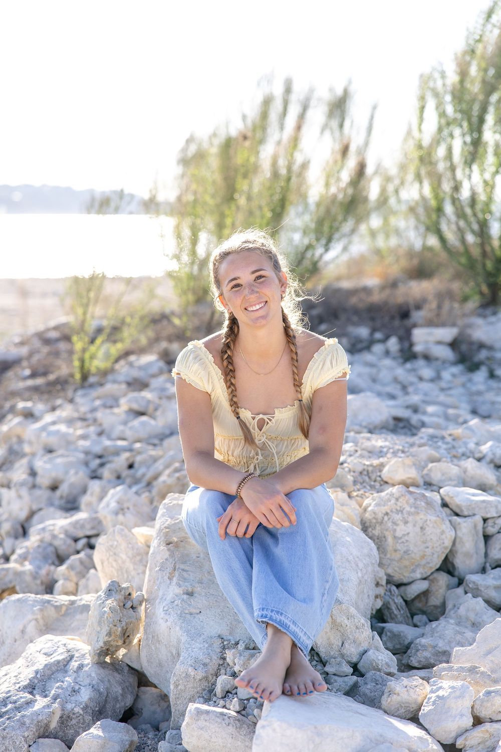 Young woman with braids sits barefoot on rocks near water, smiling in golden light, wearing yellow top and jeans.