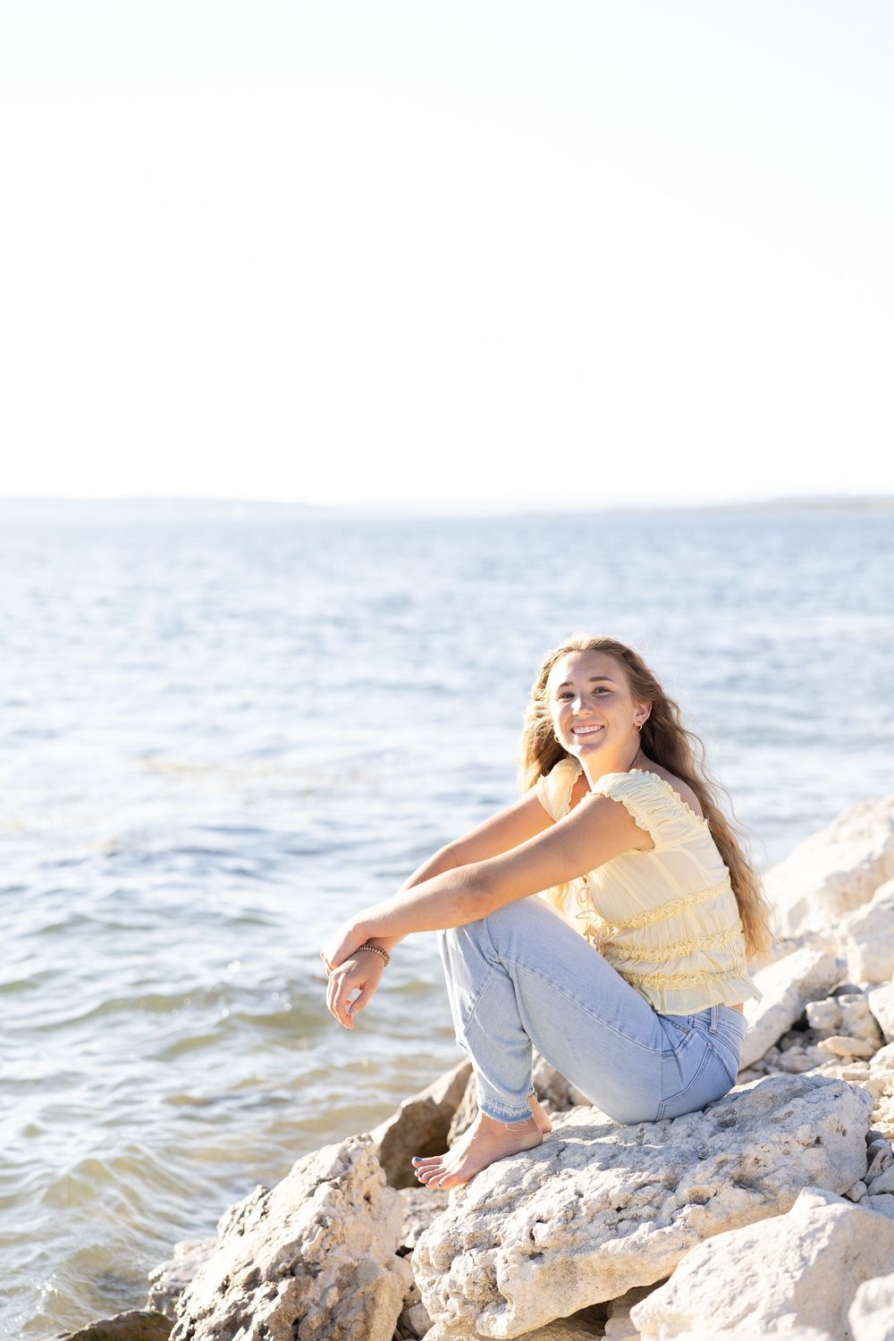 A smiling young woman with long hair sits on rocky shoreline wearing yellow top and jeans with ocean in background.