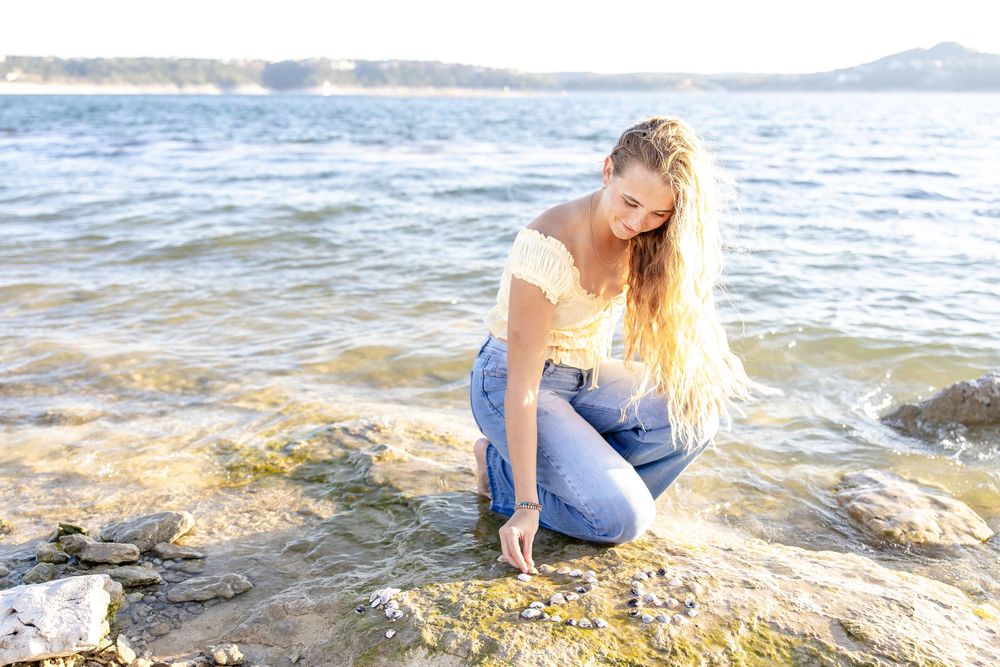 Young woman with long blonde hair crouching on rocky shoreline, touching water's edge in casual jeans and white top.