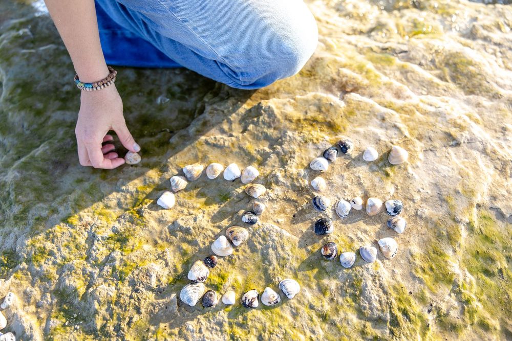 Person arranging seashells into a heart shape on a rocky beach surface near the ocean shoreline.