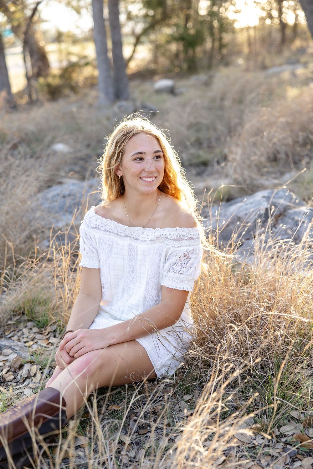 Young woman in white off-shoulder dress sitting in golden dry grass, smiling outdoors in warm sunlight.