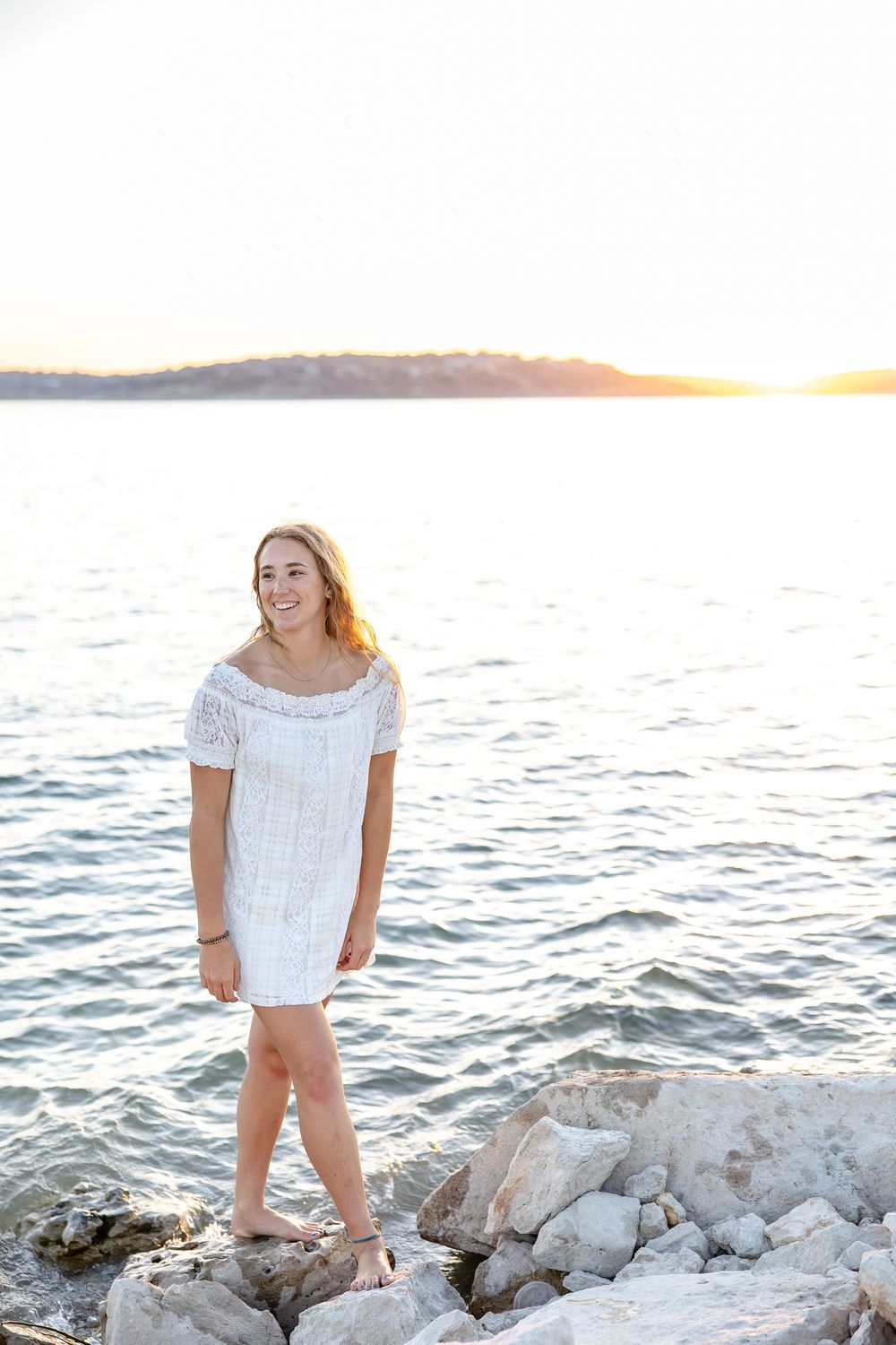 Young woman in white off-shoulder dress standing on rocks by the water at sunset.