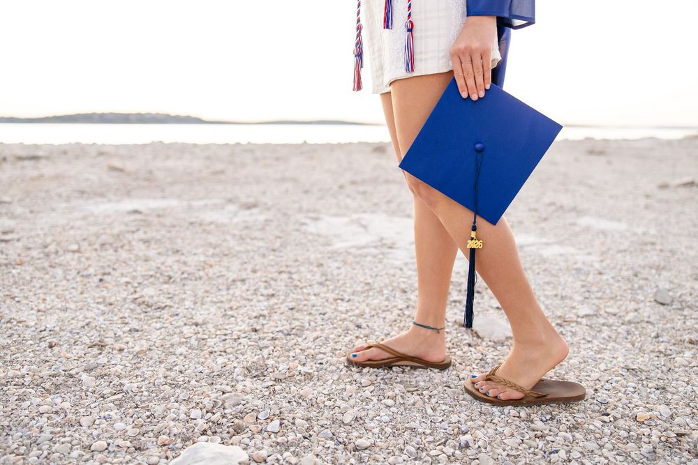 Graduate holding blue cap and diploma on rocky beach, wearing sandals and white dress.