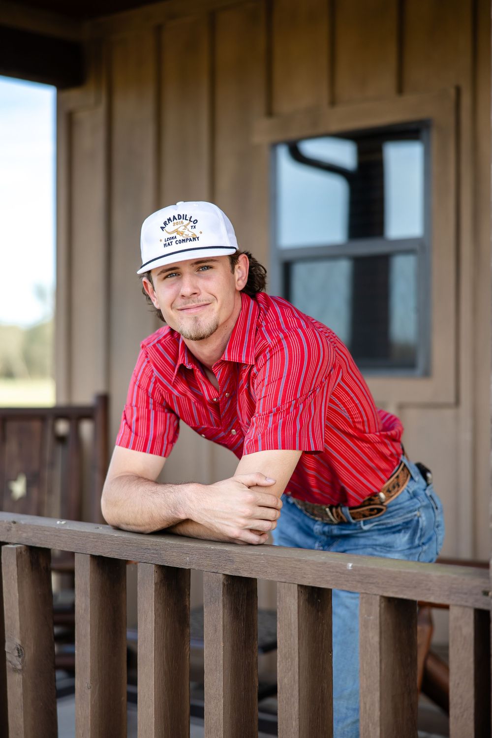 Young man in red striped shirt and white trucker hat leans on wooden porch railing, smiling outdoors.