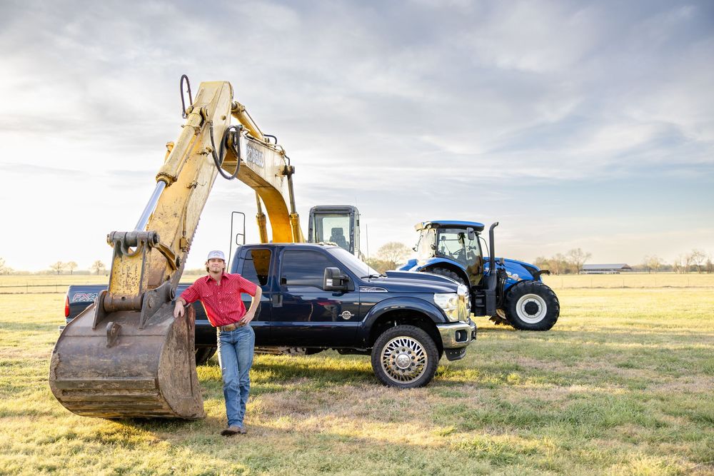Farmer in red shirt stands beside excavator, pickup truck, and blue tractor in open field at golden hour.