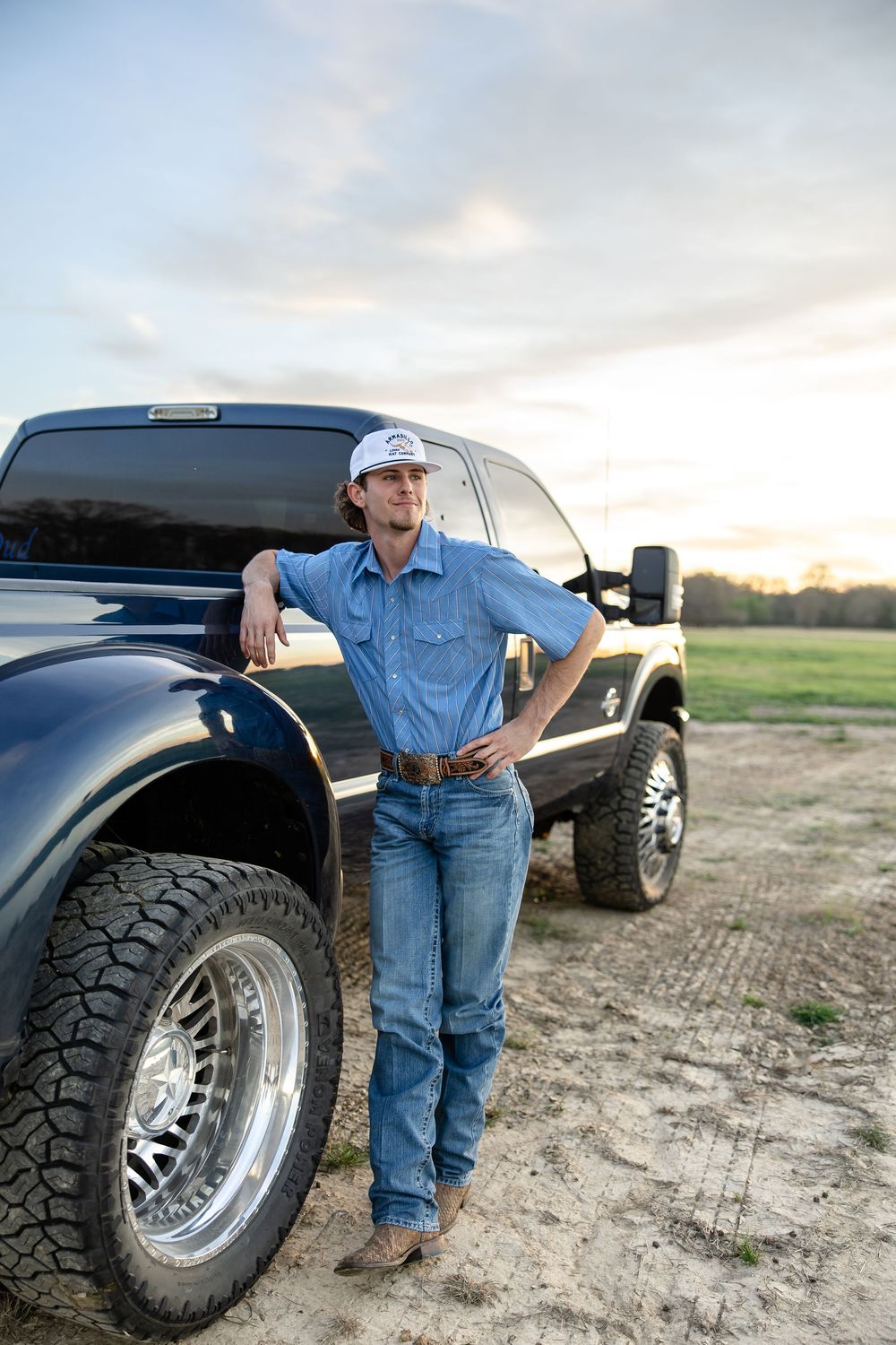 Young man in blue shirt and jeans leans against a lifted black Ford truck on a dirt road at sunset.