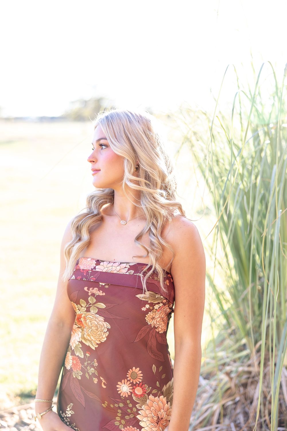 Blonde woman in floral burgundy dress standing near tall grass in golden sunlight outdoors.