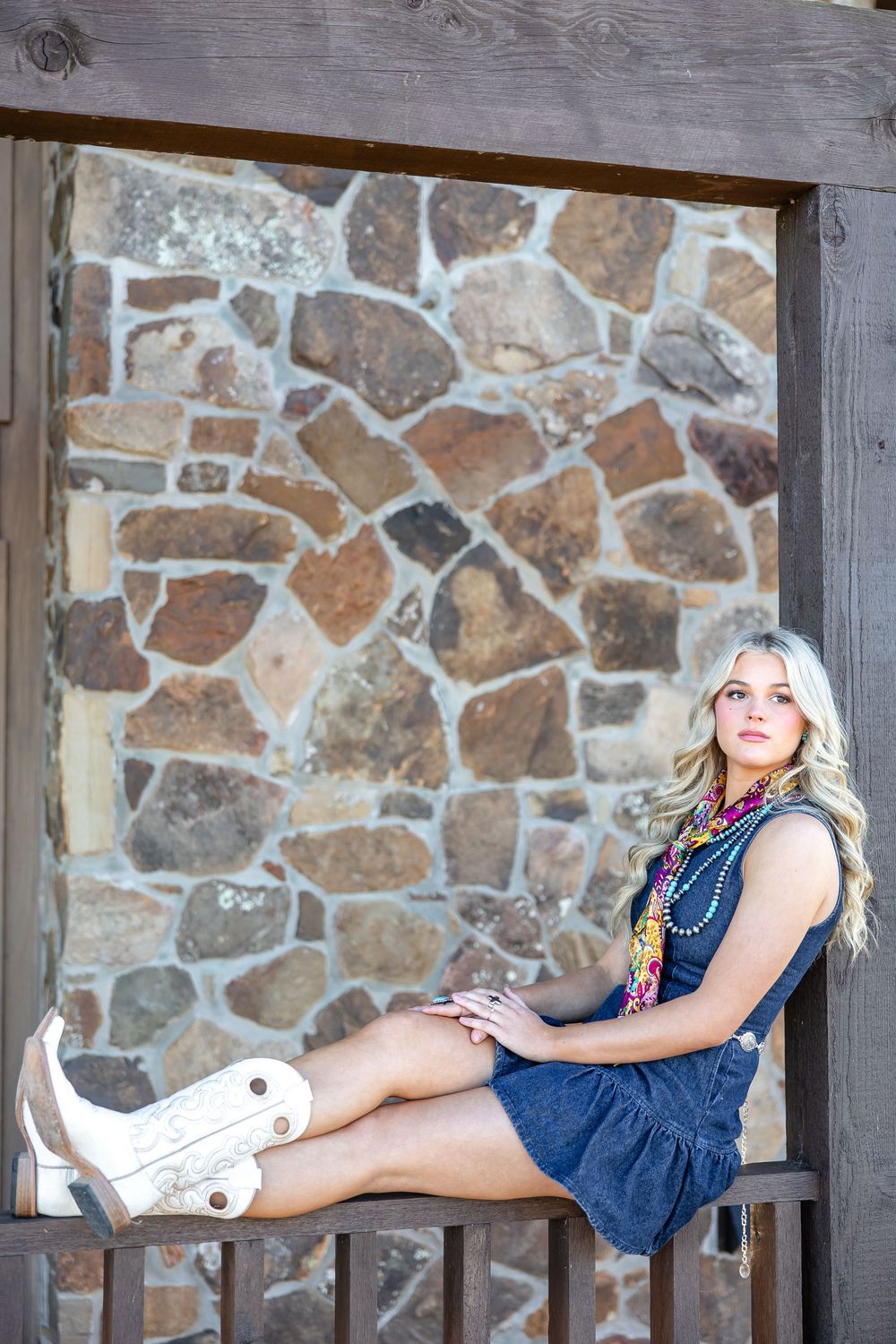 Blonde woman in denim dress and white cowboy boots sits on a fence in front of a rustic stone wall.