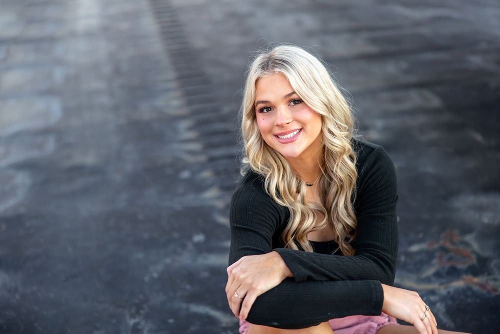 Young blonde woman smiling, wearing black top, sitting outdoors on dark pavement in a senior portrait photo.