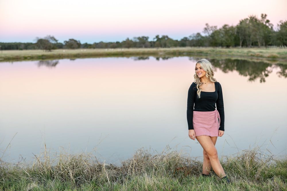 Blonde woman in black top and pink mini skirt standing by a calm reflective lake at sunset.