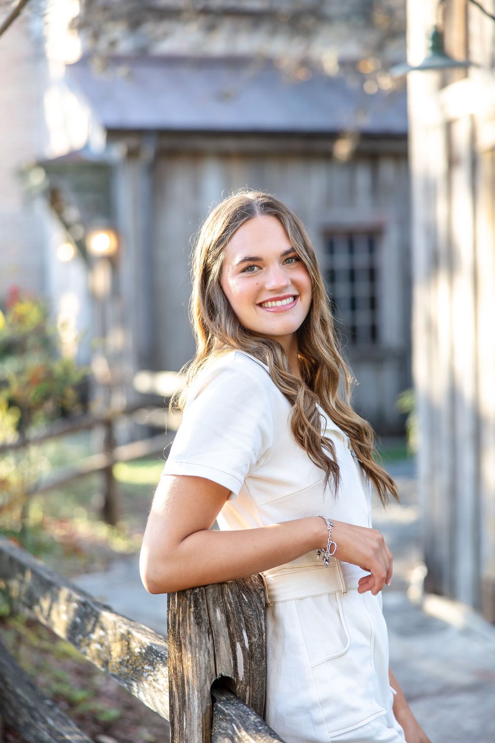 Young woman in white outfit smiling outdoors near rustic wooden fence with blurred barn background.