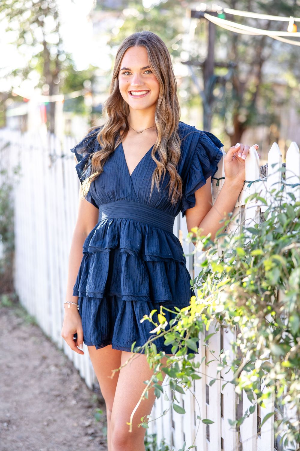 Woman in navy blue ruffle mini dress smiling by white picket fence with lush greenery.