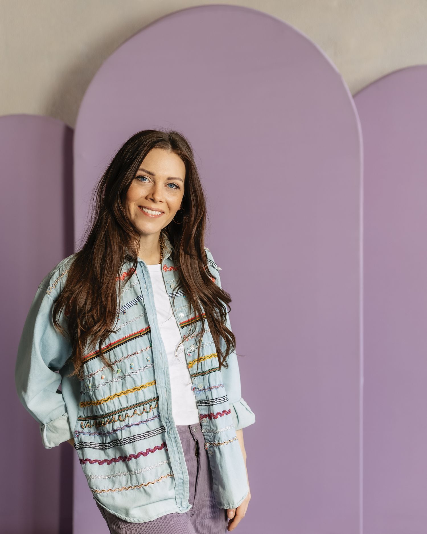 Someone with long dark hair wearing a light blue denim jacket with embroidered details poses against a purple arched wall.