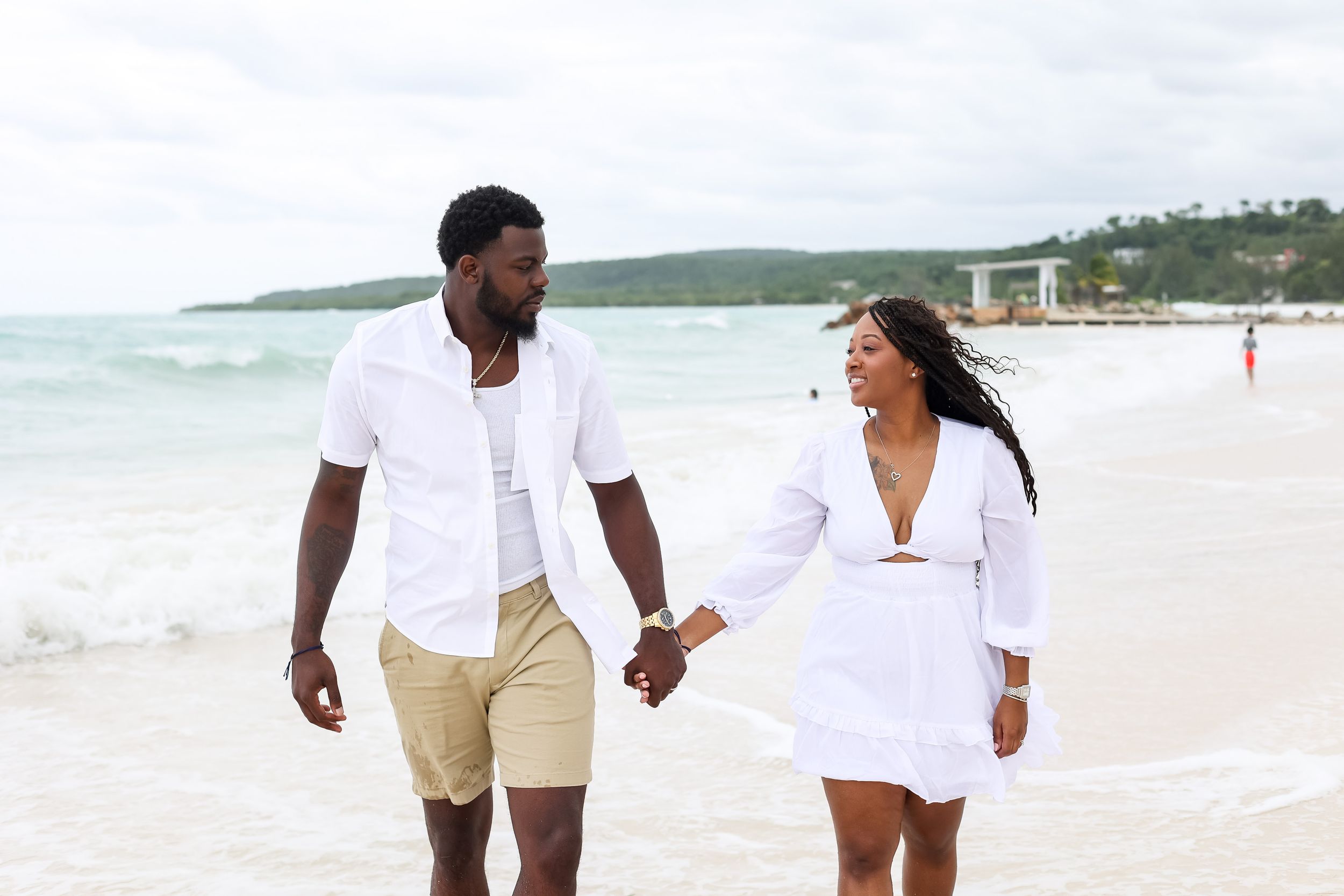 Couple holding hands walking on a tropical beach in matching white outfits.