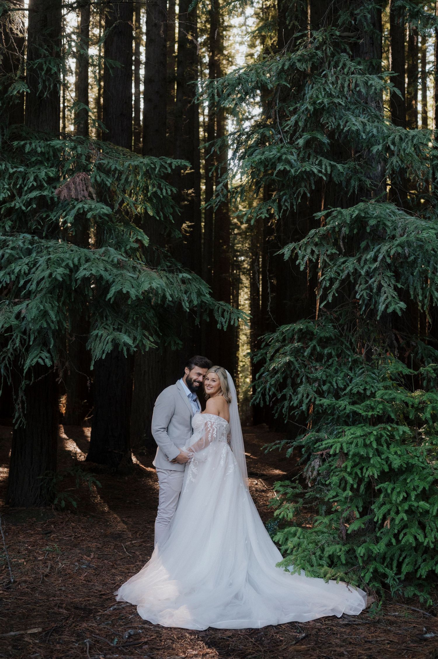 Wedding couple poses among tall evergreen trees in a forest setting with trailing white dress.
