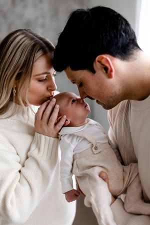 A couple in cream sweaters tenderly kiss their newborn baby, sharing a loving family moment together.