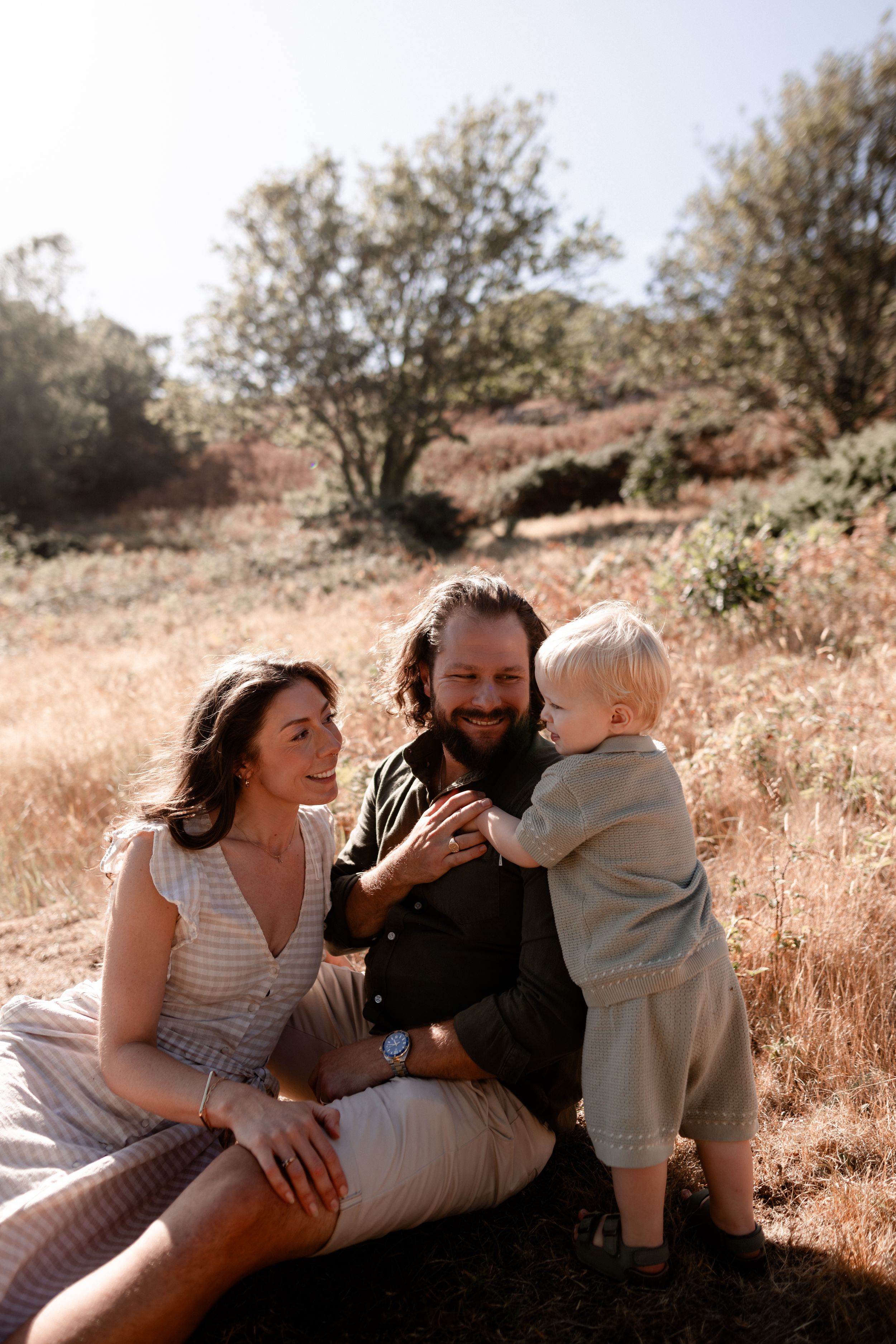 A happy family with a bearded father, brunette mother, and toddler sitting together in a sunny outdoor field.