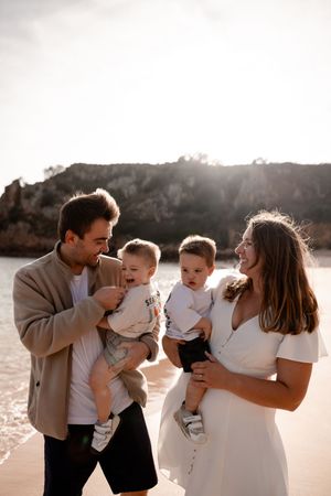 Young family with two toddlers smiling on a sunny beach, parents holding babies with rocky cliffs in the background.