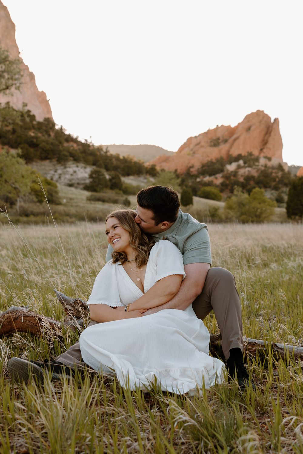 A couple shares a romantic moment sitting in tall grass at sunset with red rock formations in the background at Garden of the Gods.