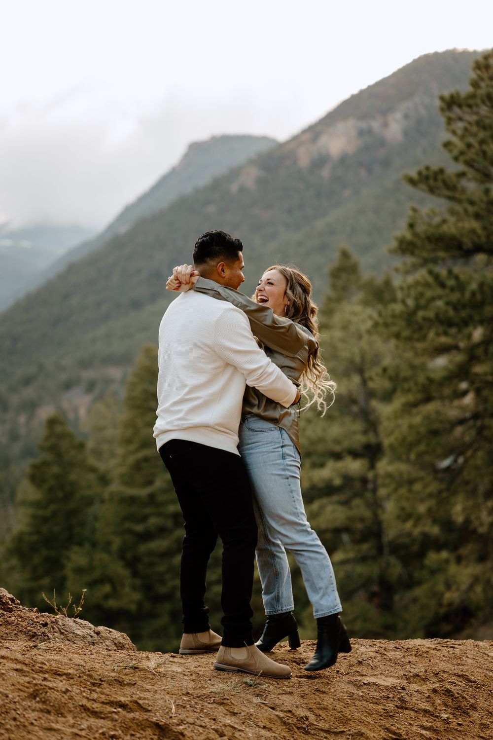 A couple embraces joyfully while standing on a rocky overlook with mountains and pine trees in the Colorado background.