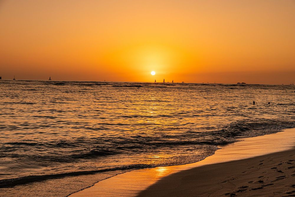 Golden sunset over ocean waves with silhouettes of people on beach and boats on horizon in Hawaii.