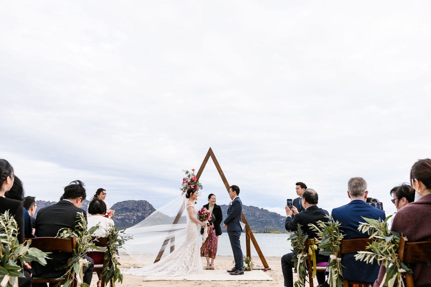A couple stands under a triangular arch during their beach wedding ceremony with mountains and ocean in the background.