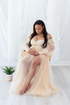 Maternity photo of woman in flowing peach gown sitting elegantly beside small potted plant.