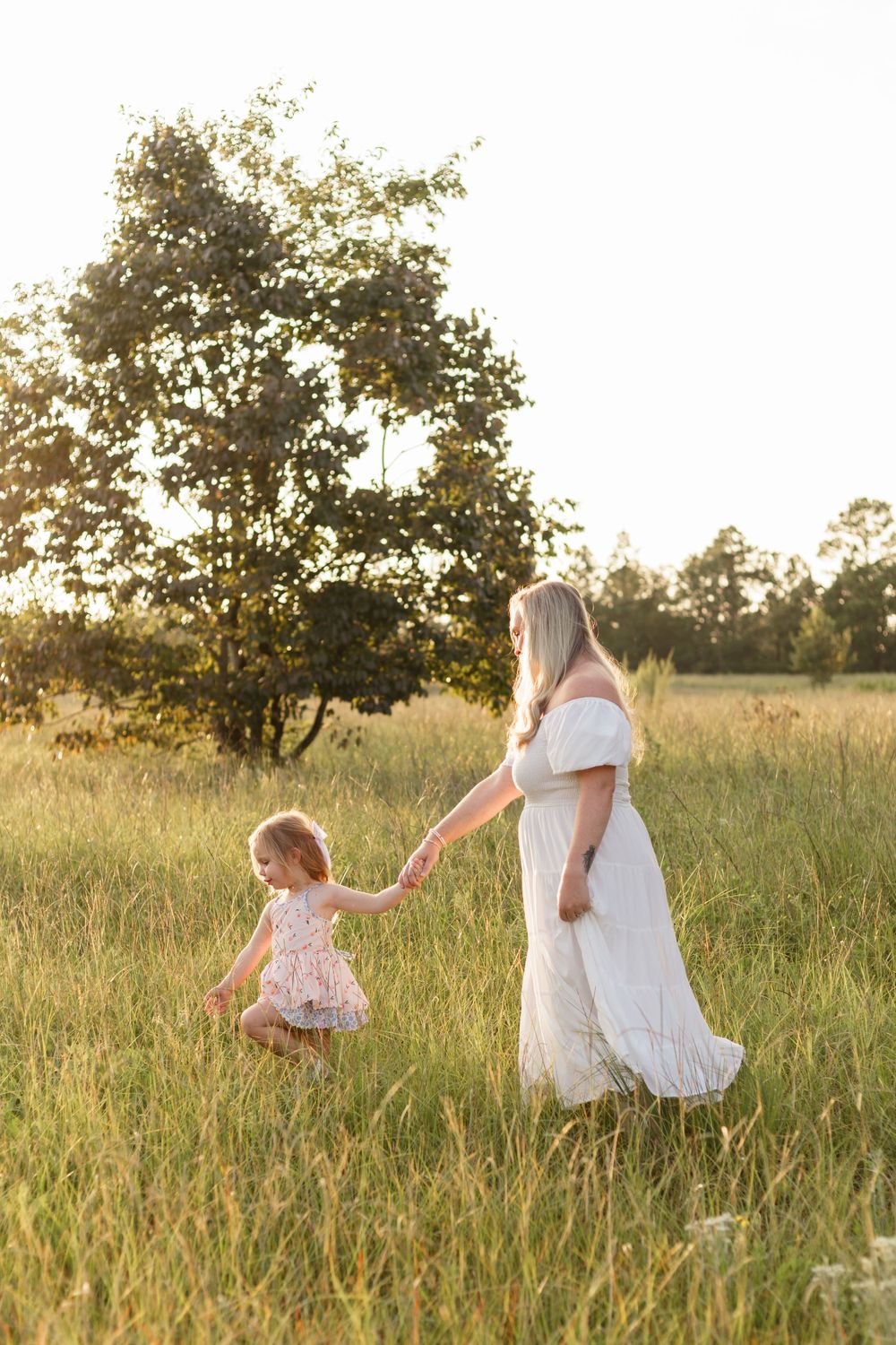 A woman in white holds hands with a small child while walking through a sunlit meadow.