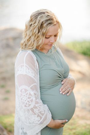 Expectant mother in a sage green dress with white lace sleeves cradles her baby bump against a blurred background.