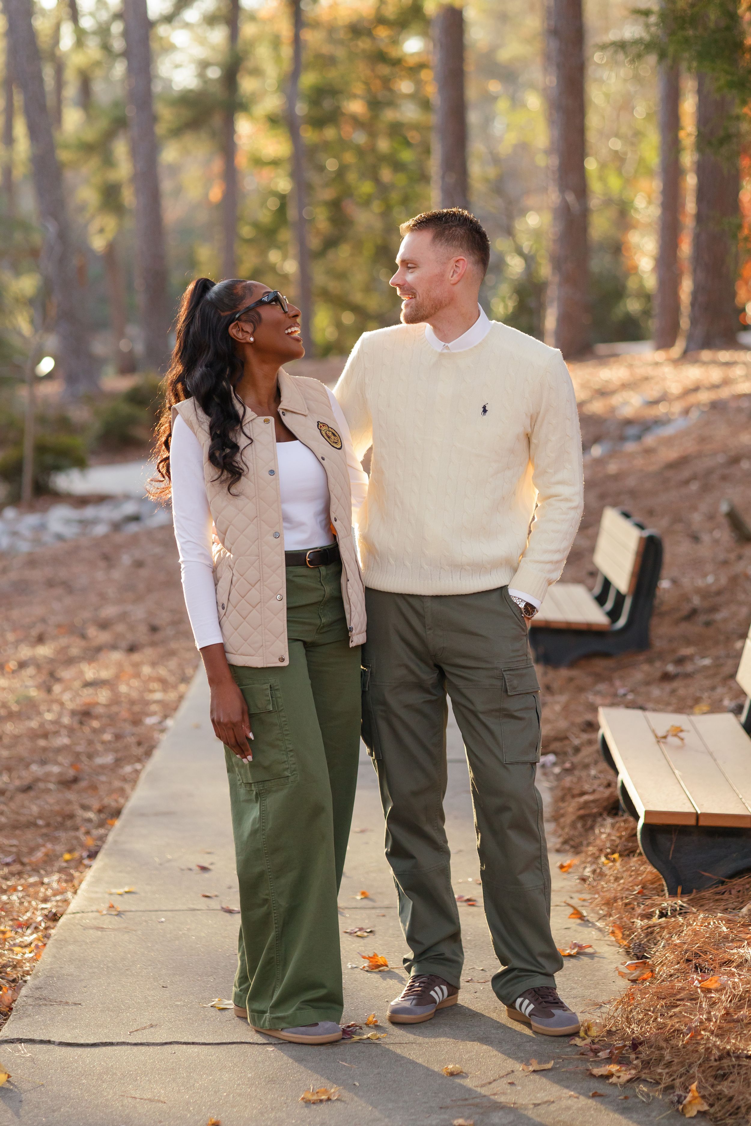 A couple in matching cream sweaters and green pants share a tender moment while walking on a wooded path at sunset.