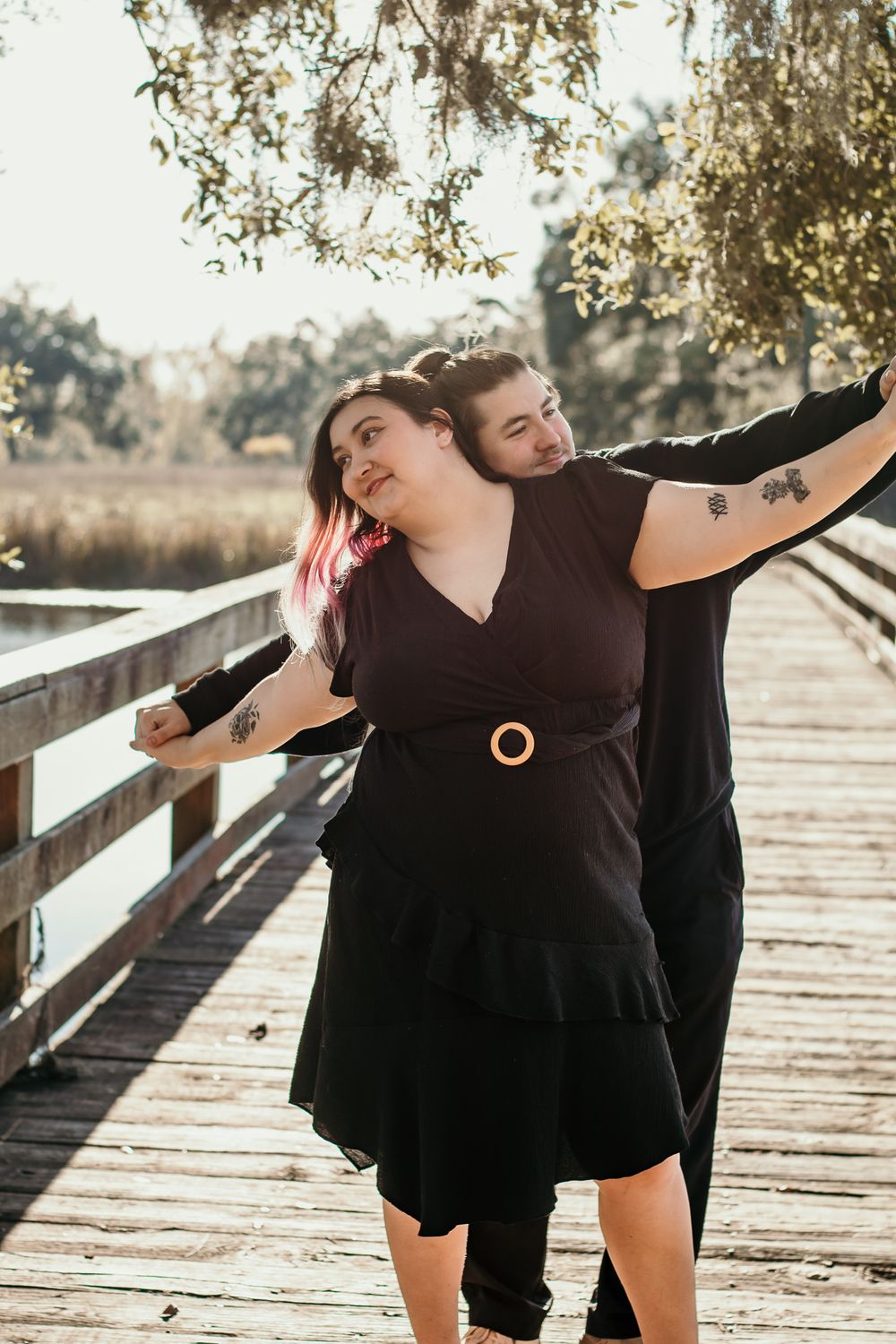 Couple in black attire striking playful pose on wooden boardwalk at sunset.