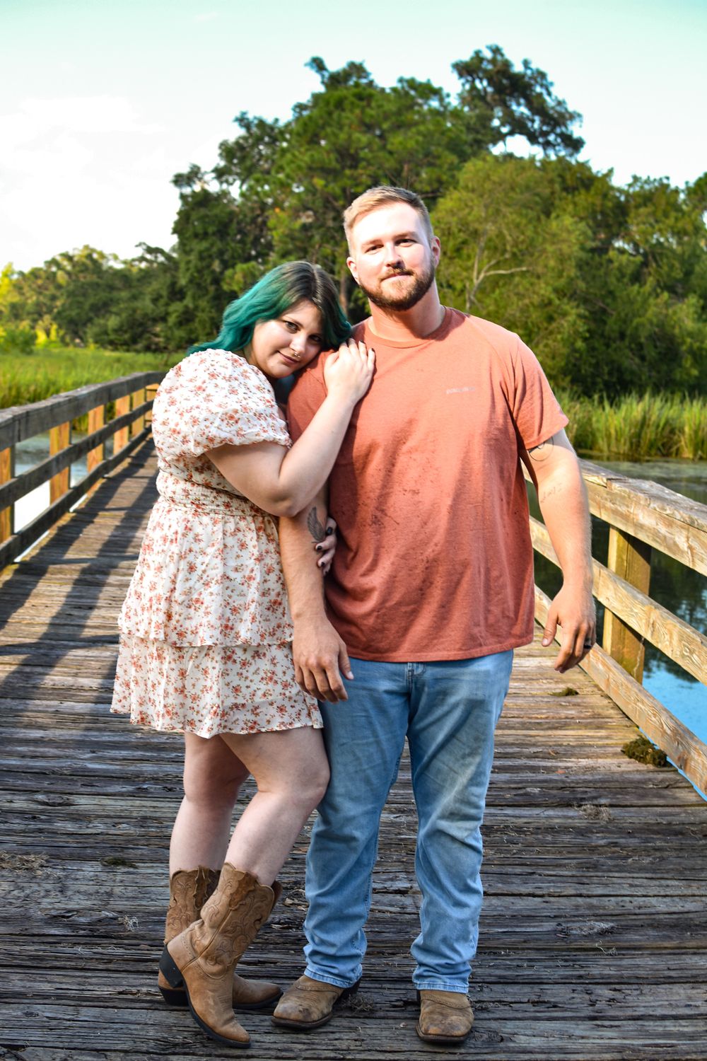 Couple posing together on wooden boardwalk with green trees in background.