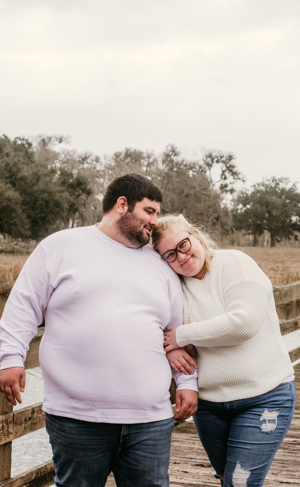 A couple wearing cozy sweaters lean against each other affectionately while standing by a wooden fence at sunset.
