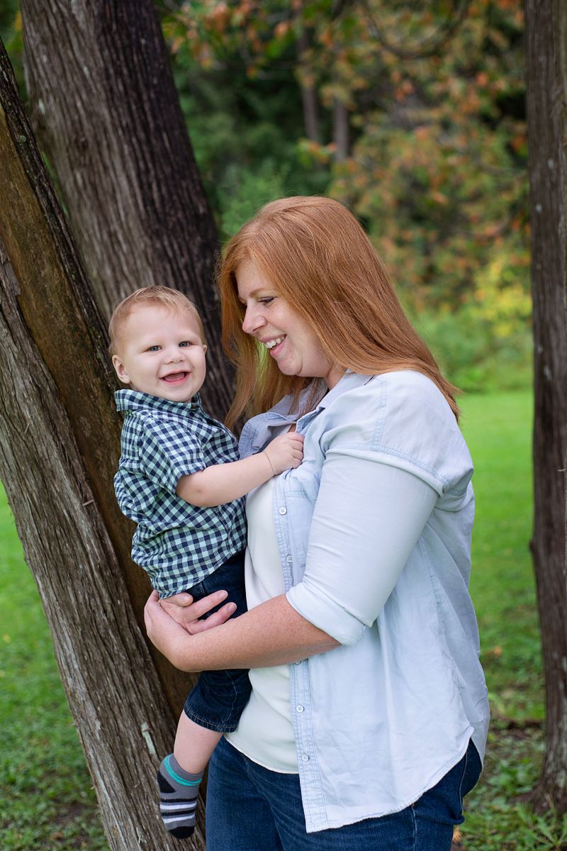 Outdoors in wooded area, mother hold toddler son and smiles at him,  while son smiles at camera