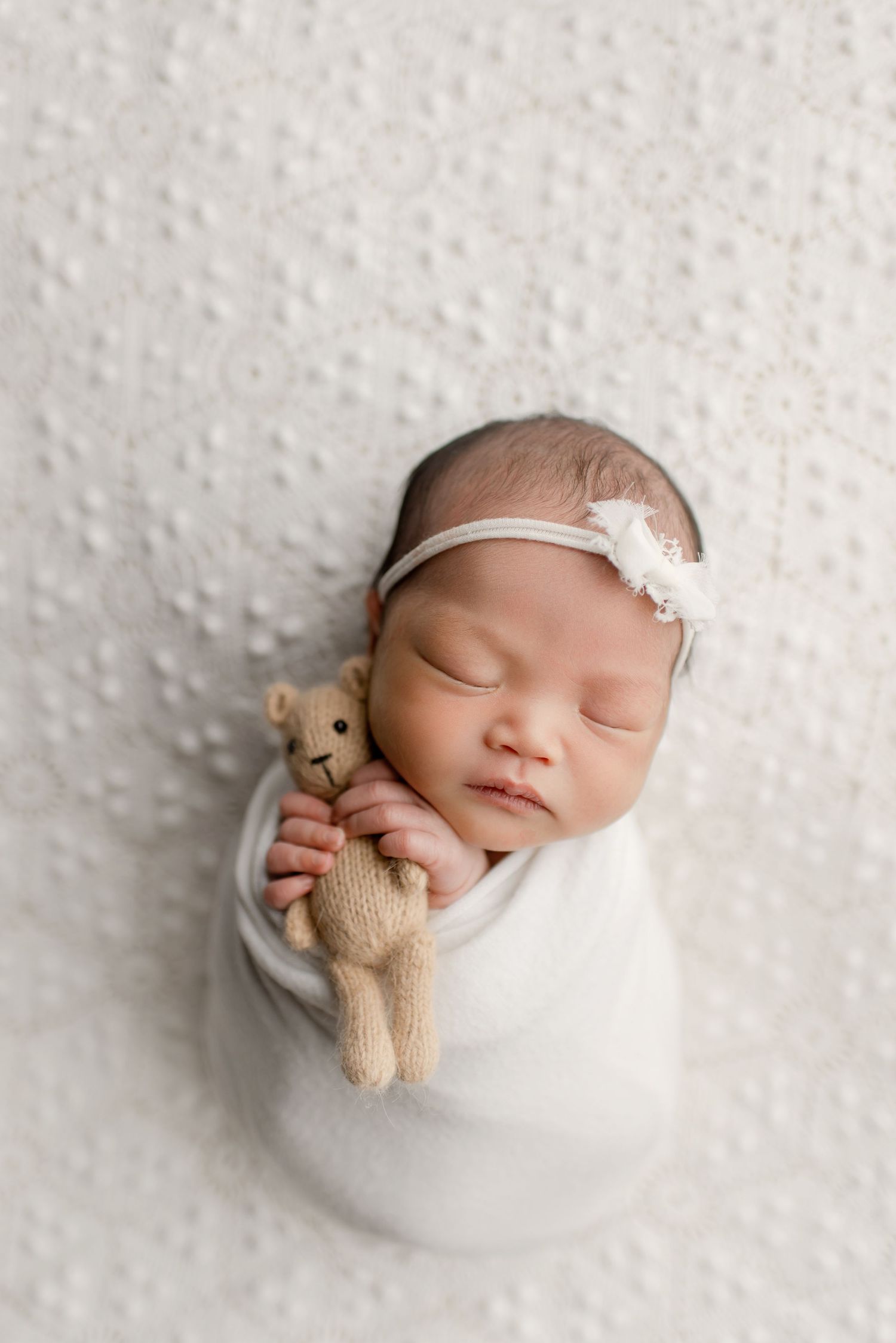 Sleeping newborn swaddled in white wrap holding tiny teddy bear with floral headband.